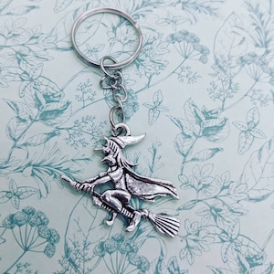 May include: A silver-toned keychain featuring a witch riding a broomstick. The witch is wearing a pointed hat and a long flowing cape. The keychain is attached to a silver key ring.