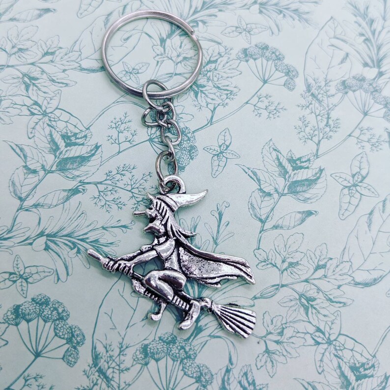 May include: A silver-toned keychain featuring a witch riding a broomstick. The witch is wearing a pointed hat and a long flowing cape. The keychain is attached to a silver key ring.