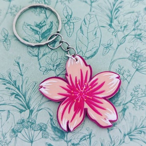 May include: A pink and white cherry blossom keychain with a silver key ring. The blossom has a pink center with yellow accents.
