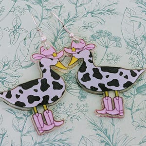 Könnte beinhalten: Ein Paar ausgefallene Ohrringe mit Entenmotiven. Jede Ente hat einen hellvioletten Körper mit schwarzen Flecken, einen rosa Cowboyhut und rosa Cowboy-Stiefel. Die Ohrringe haben silberfarbene Haken.