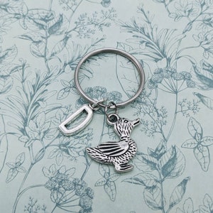 Op de afbeelding: Zilveren sleutelhanger met een eendenhanger en een letter D-hanger. De sleutelhanger bevindt zich op een achtergrond met een bloemenpatroon.