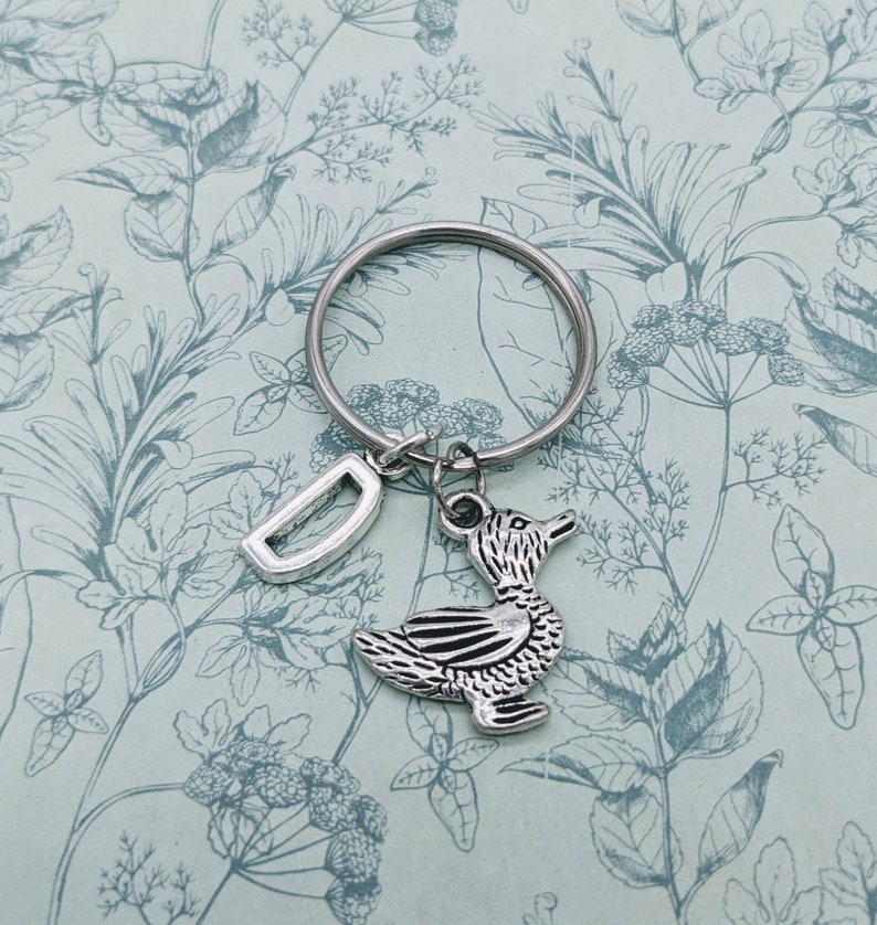 Op de afbeelding: Zilveren sleutelhanger met een eendenhanger en een letter D-hanger. De sleutelhanger bevindt zich op een achtergrond met een bloemenpatroon.