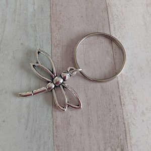 Könnte beinhalten: Silberner Libellen-Schlüsselanhänger mit einem silbernen Schlüsselring. Die Libelle hat detaillierte Flügel und einen strukturierten Körper.