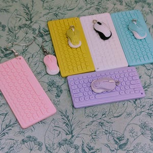 Könnte beinhalten: Eine Sammlung farbenfroher Tastatur- und Mausohrringe in Rosa, Gelb, Weiß, Blau und Lila. Jeder Ohrring hat ein Miniatur-Tastatur- und Maus-Design mit silberfarbenen Haken. Die Ohrringe sind auf einem floral gemusterten Hintergrund angeordnet.
