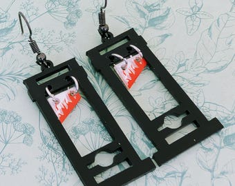 Puede incluir: Un par de pendientes de guillotina negros con cuchillas rojas y blancas. Los pendientes están hechos de metal y tienen un diseño colgante.