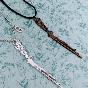 May include: Two necklaces with gun-shaped pendants. One necklace has a silver pendant and the other has a bronze pendant. Both pendants are detailed with a textured design.