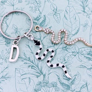 May include: A silver key chain with a letter D charm, a black and white snake charm, and a clear rhinestone snake charm.