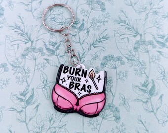 May include: A novelty keychain featuring a pink bra design with the text "BURN YOUR BRAS" in black and white. A lit match is also included in the design. The keychain has a silver metal ring and chain.