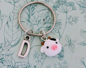 May include: A silver keychain with a large ring, a letter "D" charm, and a white cow charm with black spots and brown horns. The cow has a pink nose and is smiling. The background is a light green floral pattern.