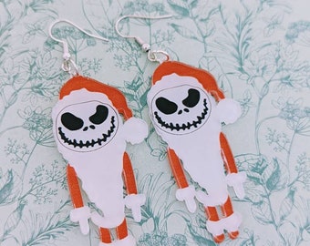 May include: A pair of dangle earrings featuring a white Jack Skellington character wearing a red Santa hat. The earrings are made of acrylic and have silver hooks.