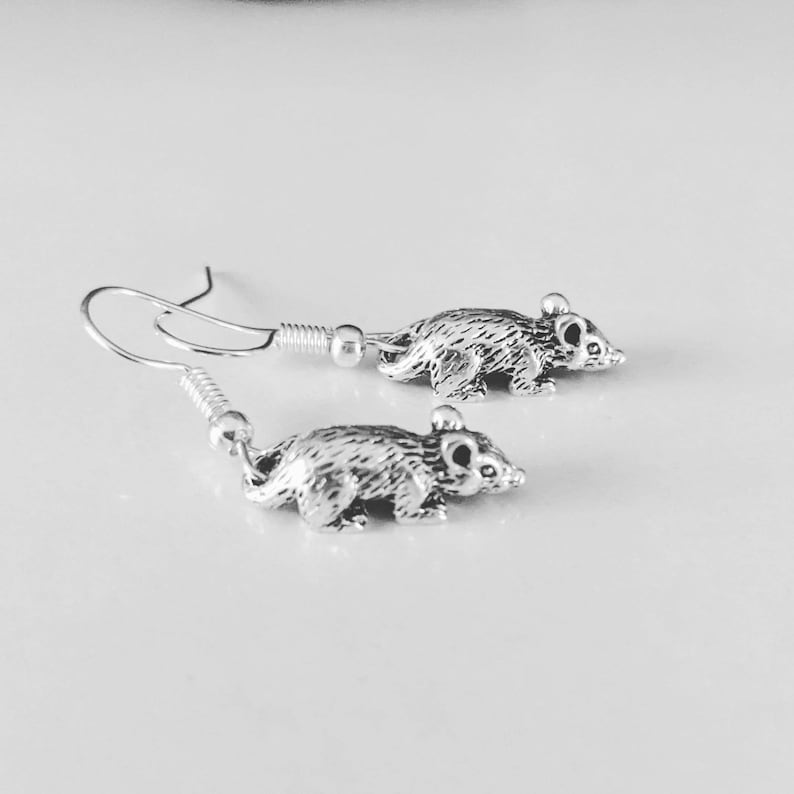 May include: A pair of silver earrings featuring a small, detailed rat charm. The earrings have a simple hook design.