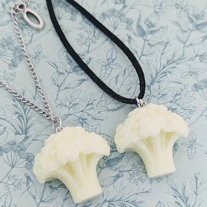 May include: Two necklaces featuring cauliflower pendants. One pendant is on a silver chain, the other on a black cord. The off-white pendants are detailed, mimicking the vegetable. The background has a floral pattern.