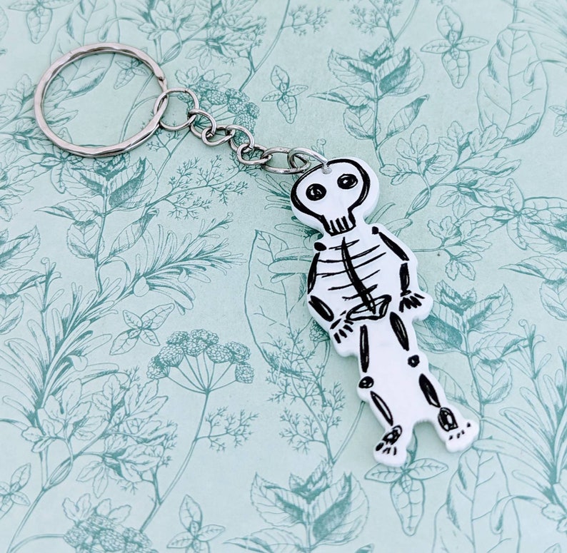 Pode incluir: Um chaveiro de esqueleto branco com um anel de chave prateado. O esqueleto &eacute; desenhado em preto e tem uma caveira com dois olhos pretos.