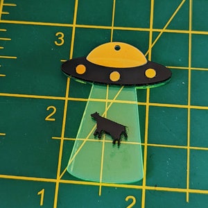 May include: A black and yellow UFO with a black silhouette of a cow underneath. The UFO is made of acrylic and has a green glow.