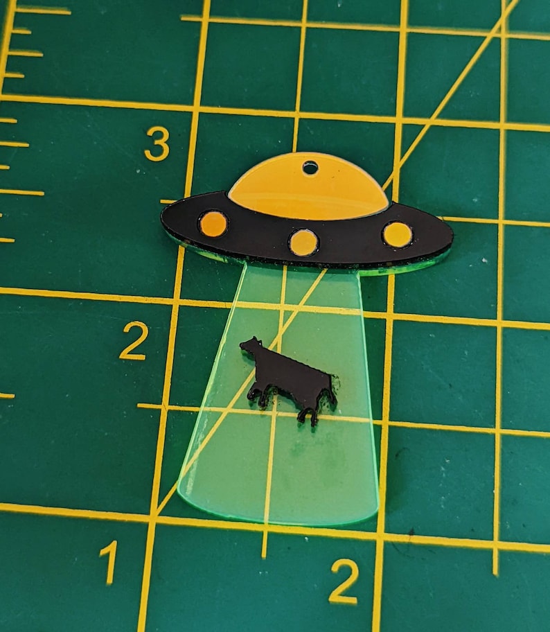 May include: A black and yellow UFO with a black silhouette of a cow underneath. The UFO is made of acrylic and has a green glow.