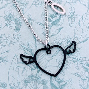 May include: A silver chain necklace with a black heart pendant with wings. The heart is open and the wings are on the side. There is a small silver charm on the chain.