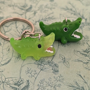 May include: Two green alligator keychains with open mouths and white teeth rest on a patterned background. The keychains are made of a glossy material and have black eyes. One keychain is attached to a silver keyring.