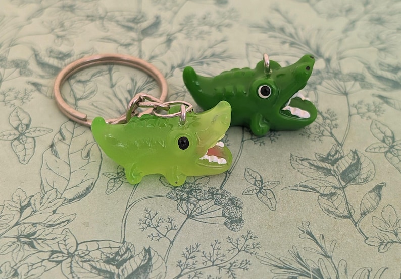 May include: Two green alligator keychains with open mouths and white teeth rest on a patterned background. The keychains are made of a glossy material and have black eyes. One keychain is attached to a silver keyring.