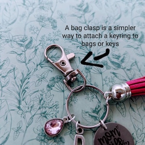 Puede incluir: Un cierre de bolso plateado con un llavero que sostiene dijes. Los dijes incluyen una gema rosa en forma de l&aacute;grima, un dije con la letra "D" y un dije redondo con las palabras "mom wife boss". Tambi&eacute;n hay una borla rosa adjunta. El texto de la imagen dice: "Un cierre de bolso es una forma m&aacute;s sencilla de sujetar un llavero a bolsos o llaves."