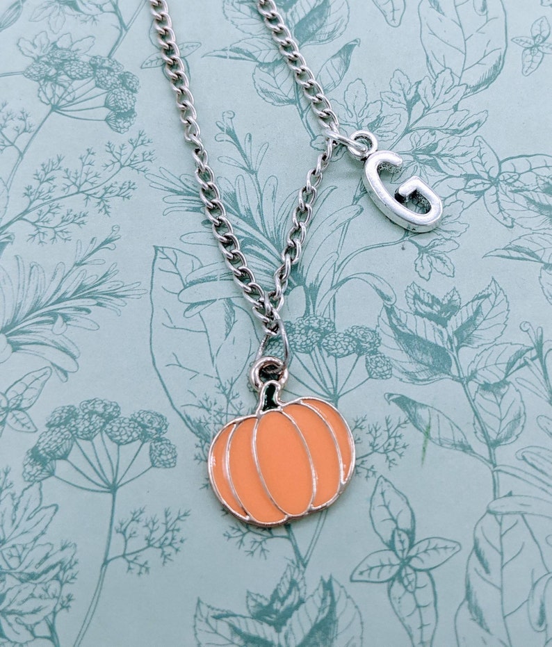 May include: A silver chain necklace with a small orange pumpkin charm and a silver charm with the letter "G" hanging from it.