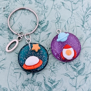 Peut inclure: Un porte-clés argenté avec un charme en forme de lettre O et deux porte-clés ronds, pailletés, sur le thème de l'espace. Un porte-clés est violet avec un vaisseau spatial bleu et une fusée avec un œuf frit. L'autre porte-clés est vert avec une étoile jaune et un vaisseau spatial avec un œuf blanc.