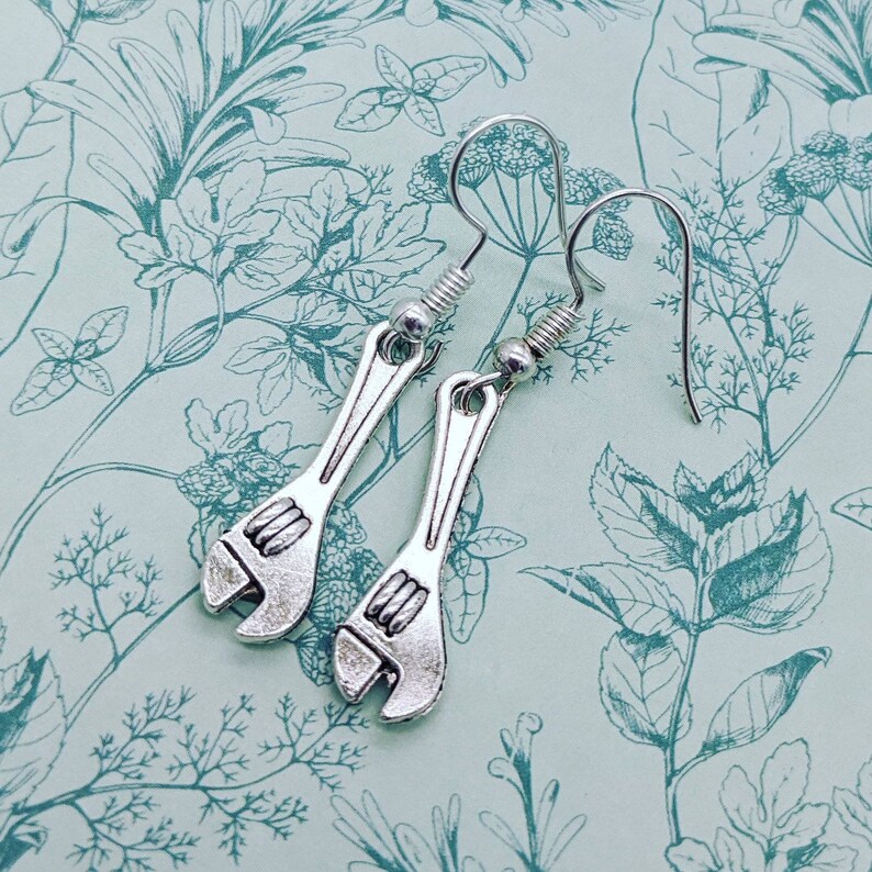 May include: Silver wrench earrings with a hook closure. The earrings are on a light green background with a floral pattern.