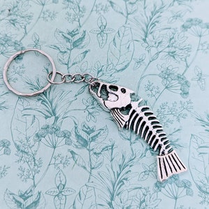 May include: Silver fish skeleton keychain with a silver key ring. The keychain is on a light blue background with a floral pattern.