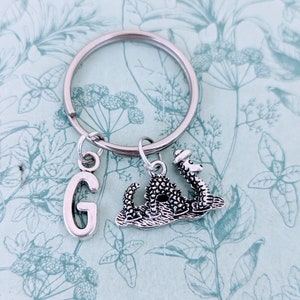 May include: A silver keychain with a letter G charm and a small silver dragon charm. The dragon charm is wearing a hat.