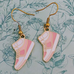 May include: Gold-tone drop earrings with a sneaker design. The sneakers are white, light pink, and dark pink with gold-tone details. The earrings hang from gold-tone ear wires.