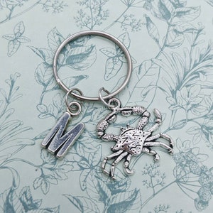 May include: Silver keychain with a letter M charm and a crab charm. The keychain is on a white background with a floral pattern.