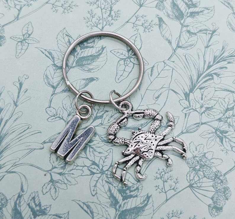 May include: Silver keychain with a letter M charm and a crab charm. The keychain is on a white background with a floral pattern.