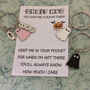 May include: Keychains with ghost charms in pink, white, and black, attached to silver keyrings. A card with the text "GHOST HUG" and a message about caring is in the background. The charms have a Halloween theme.
