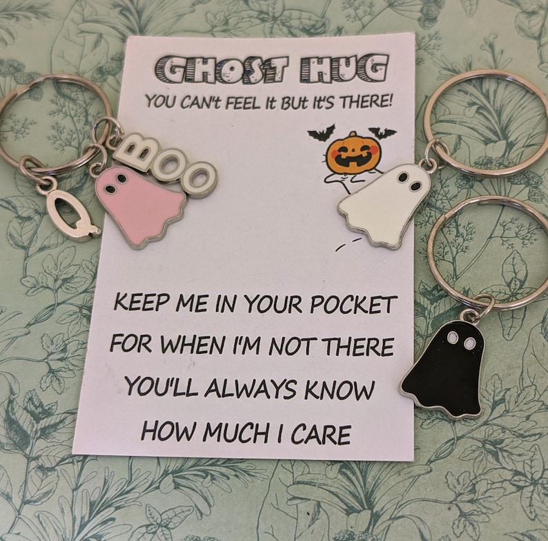 May include: Keychains with ghost charms in pink, white, and black, attached to silver keyrings. A card with the text "GHOST HUG" and a message about caring is in the background. The charms have a Halloween theme.
