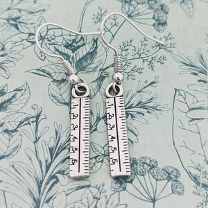 May include: Silver earrings with a ruler charm. The ruler charm has numbers 1 to 6 printed on it.