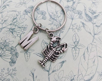 May include: Silver lobster charm keychain with a silver letter M charm. The keychain is on a white background with a floral pattern.
