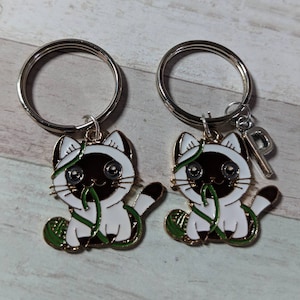 May include: Two silver keychains with a cat charm. Each cat is white with black markings and is holding a green yarn ball. One keychain has a silver charm with the letter "P" on it.