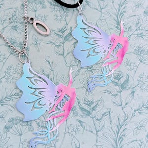 May include: Two butterfly wing pendants with a dancer silhouette, one on a silver chain and the other on a black cord. The wings are a gradient of blue and pink, with intricate cut-out designs. The background is a floral pattern.