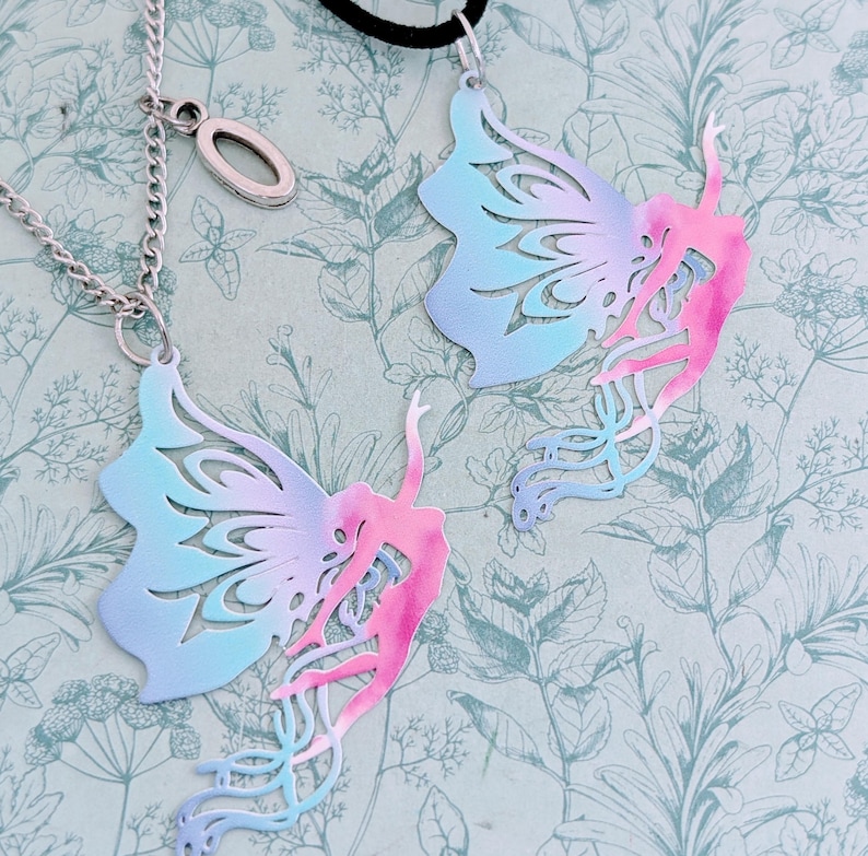 May include: Two butterfly wing pendants with a dancer silhouette, one on a silver chain and the other on a black cord. The wings are a gradient of blue and pink, with intricate cut-out designs. The background is a floral pattern.
