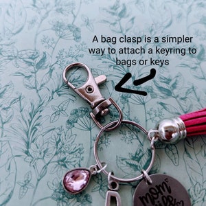 May include: A silver keyring with a pink crystal charm, a silver letter 'D' charm, and a silver charm with the text 'mom wife boss' on it. The keyring is attached to a red tassel.