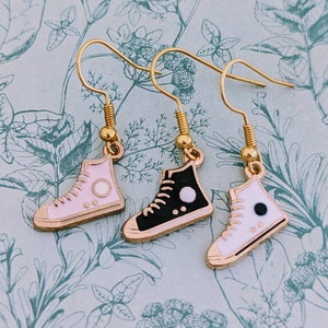 Puede incluir: Tres pares de pendientes de oro con diseño de zapatilla. Un par es rosa con detalles blancos, un par es negro con detalles blancos y un par es blanco con detalles negros.