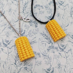 Könnte beinhalten: Zwei Halsketten mit Mais-Anhängern. Eine Halskette hat eine silberne Kette und einen silbernen Anhänger mit der Aufschrift "G". Die andere Halskette hat eine schwarze Schnur und einen gelben Mais-Anhänger.