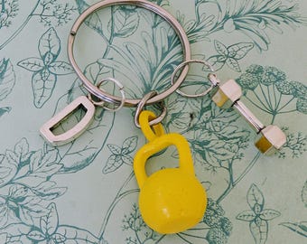 May include: A silver keyring with three charms: a silver letter "D", a yellow kettlebell, and a silver dumbbell. The charms are attached to a large silver ring. The background is a light green paper with a floral pattern.