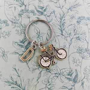 May include: A silver keyring with a gold-toned bicycle charm and a silver letter "D" charm. The bicycle charm has black detailing and white wheels. The background features a light green floral pattern.