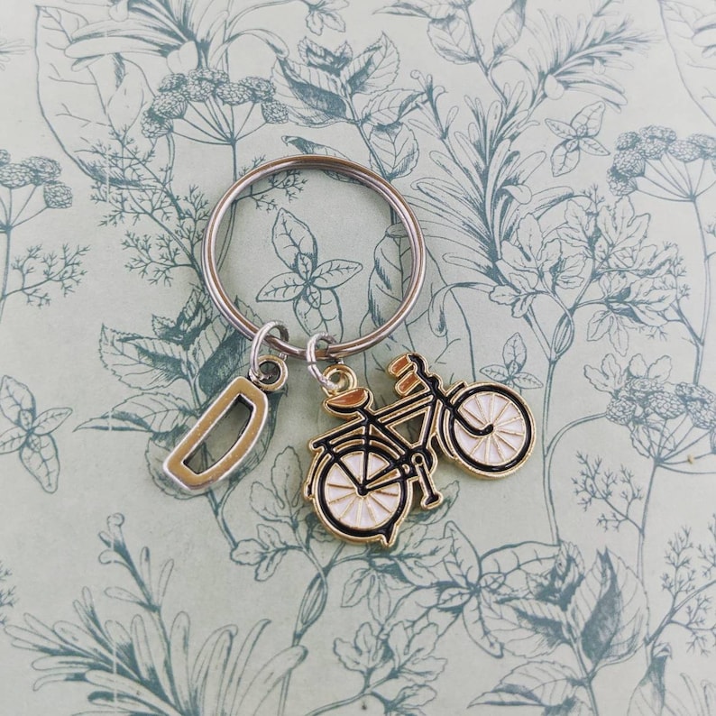 May include: A silver keyring with a gold-toned bicycle charm and a silver letter "D" charm. The bicycle charm has black detailing and white wheels. The background features a light green floral pattern.