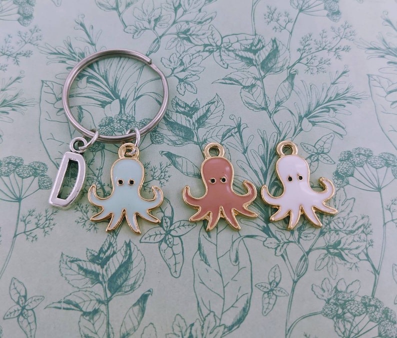 May include: A silver keychain with a letter "D" charm and three octopus charms in light blue, pink, and white. The octopus charms have gold-colored outlines and black eyes. The background has a floral pattern.