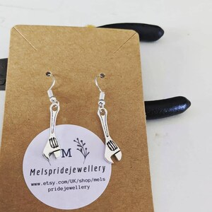 May include: Silver wrench earrings on a brown card with the text "Melspridejewellery" and "www.etsy.com/UK/shop/mels pridejewellery".