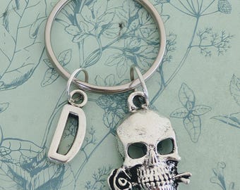 May include: Silver-toned keychain featuring a skull charm with a rose and a letter "D" charm. The skull has detailed features and is attached to a large metal ring. The background is a light blue floral pattern.