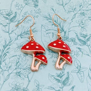 May include: A pair of gold-tone earrings featuring red mushroom charms with white polka dots. The mushrooms have a three-dimensional design and are connected to the earrings with a gold-tone loop.