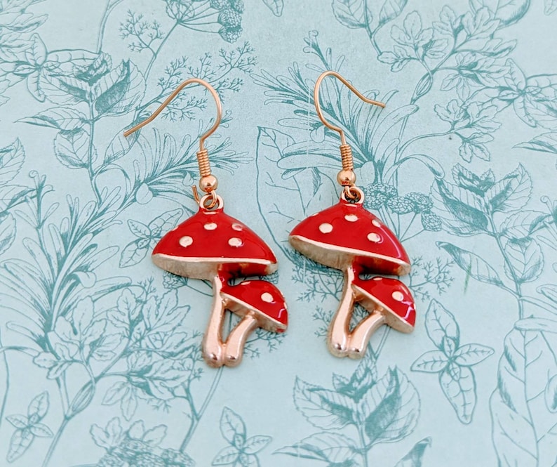 May include: A pair of gold-tone earrings featuring red mushroom charms with white polka dots. The mushrooms have a three-dimensional design and are connected to the earrings with a gold-tone loop.