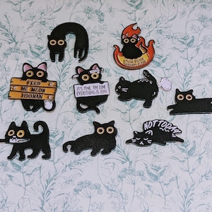 May include: A collection of black cat-shaped stickers with various designs and text. Some cats have signs that read "FEED ME MEOW HOOMAN" and "IT'S FINE, I'M FINE EVERYTHING IS FINE". Other stickers feature cats in flames and the words "NOT TODAY".